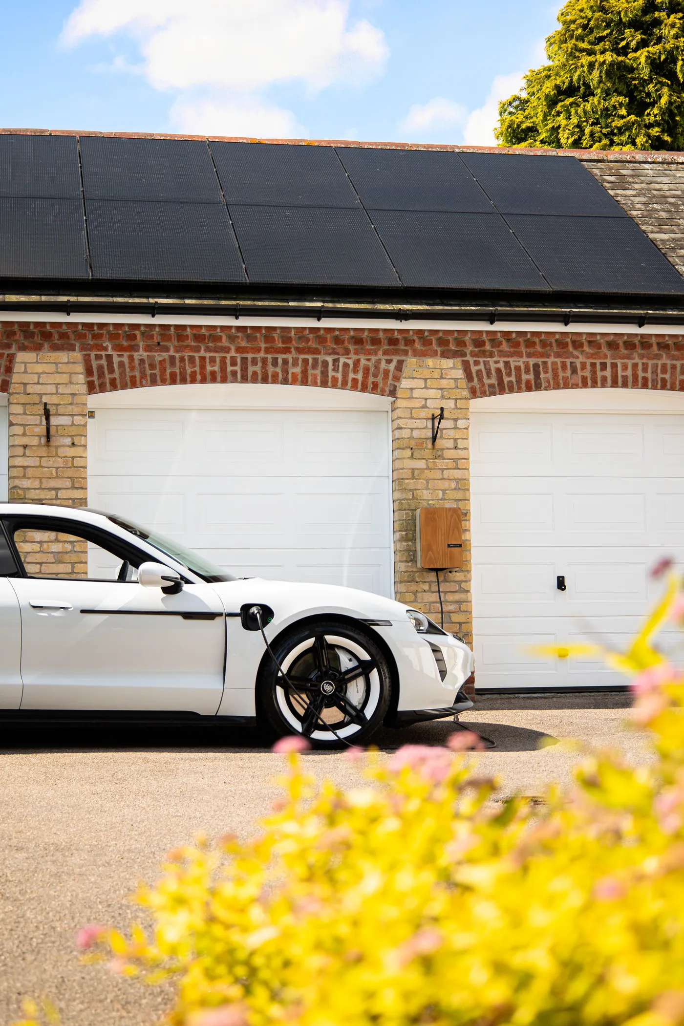 An electric car charging from a wall-mounted home charger beside garages with a solar roof